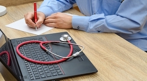 Médico escrevendo num papel e um estetoscópio sobre o laptop